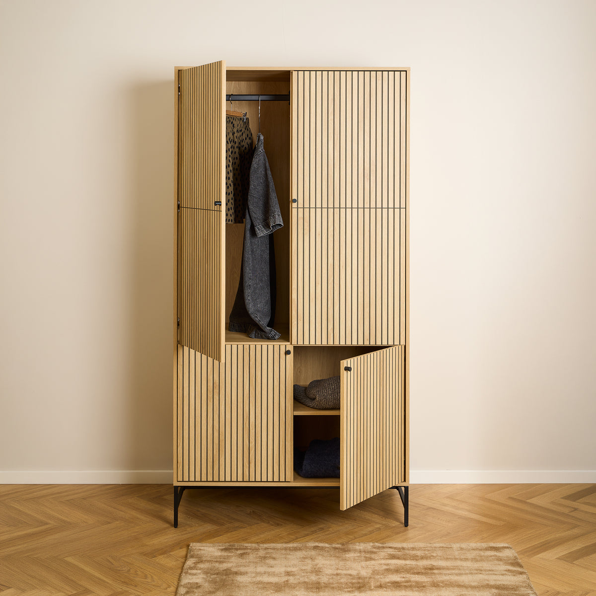 Armoire Bambouz 99 x 200 posée à même le sol, avec un motif en chevrons – portes ouvertes, vêtements suspendus à une tringle et pulls rangés sur une étagère, armoire de chambre à coucher