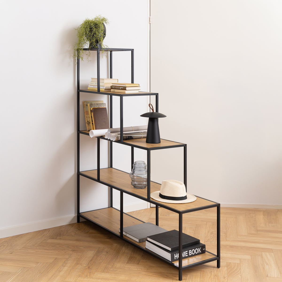 Étagère Skeleto 135x150 Chêne Sauvage dans le coin de la pièce sur un parquet à chevrons, avec des livres, une lampe et un chapeau – étagère en escalier