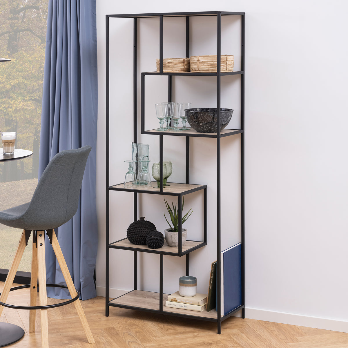 Étagère Skeleto 77 x 185, chêne Sonoma, près d'une fenêtre ornée d'un rideau bleu, à côté d'une chaise – étagère pour livres et verres, pour le salon