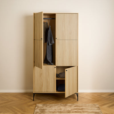 Armoire Bambouz 99 x 200 posée à même le sol, avec un motif en chevrons – portes ouvertes, vêtements suspendus à une tringle et pulls rangés sur une étagère, armoire de chambre à coucher
